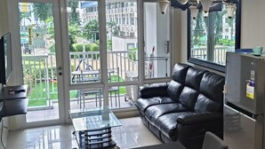 Un salon moderne avec un canapé en cuir noir, une table basse en verre et une table à manger avec des chaises. Il y a un téléviseur sur le côté gauche et un réfrigérateur sur le côté droit. La pièce est dotée de grandes fenêtres qui offrent une vue sur un balcon et sur la verdure à l’extérieur.