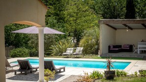 A pool area with lounge chairs, a covered seating area, and a garden with rocks and plants. - One-storey villa with air conditioning and pool (Bagnols en Forêt)