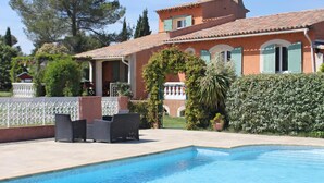 A swimming pool with a tiled deck, a patio area with outdoor furniture, and a large house with a tiled roof and green shutters. - Child-friendly holiday home with pool (Bagnols en Forêt)