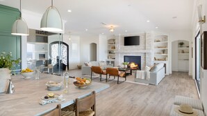 A spacious living area with a fireplace, a dining table set with fruits and glasses, and a modern kitchen in the background. - PInot Point by AvantStay | Valley Estate Mins to Wineries | Pool + Home Theater (Temecula)