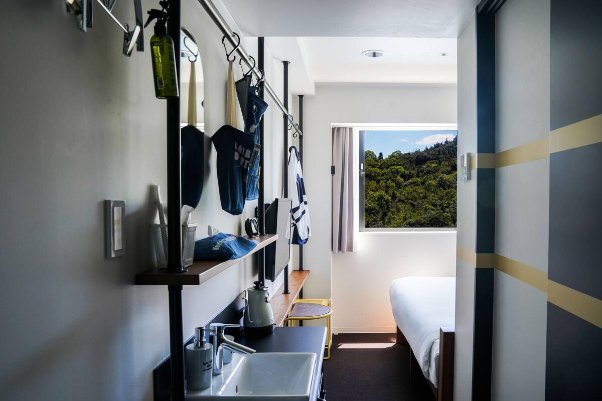 A compact hotel room with a bed, a small table, a sink, and a window offering a view of a forested area.