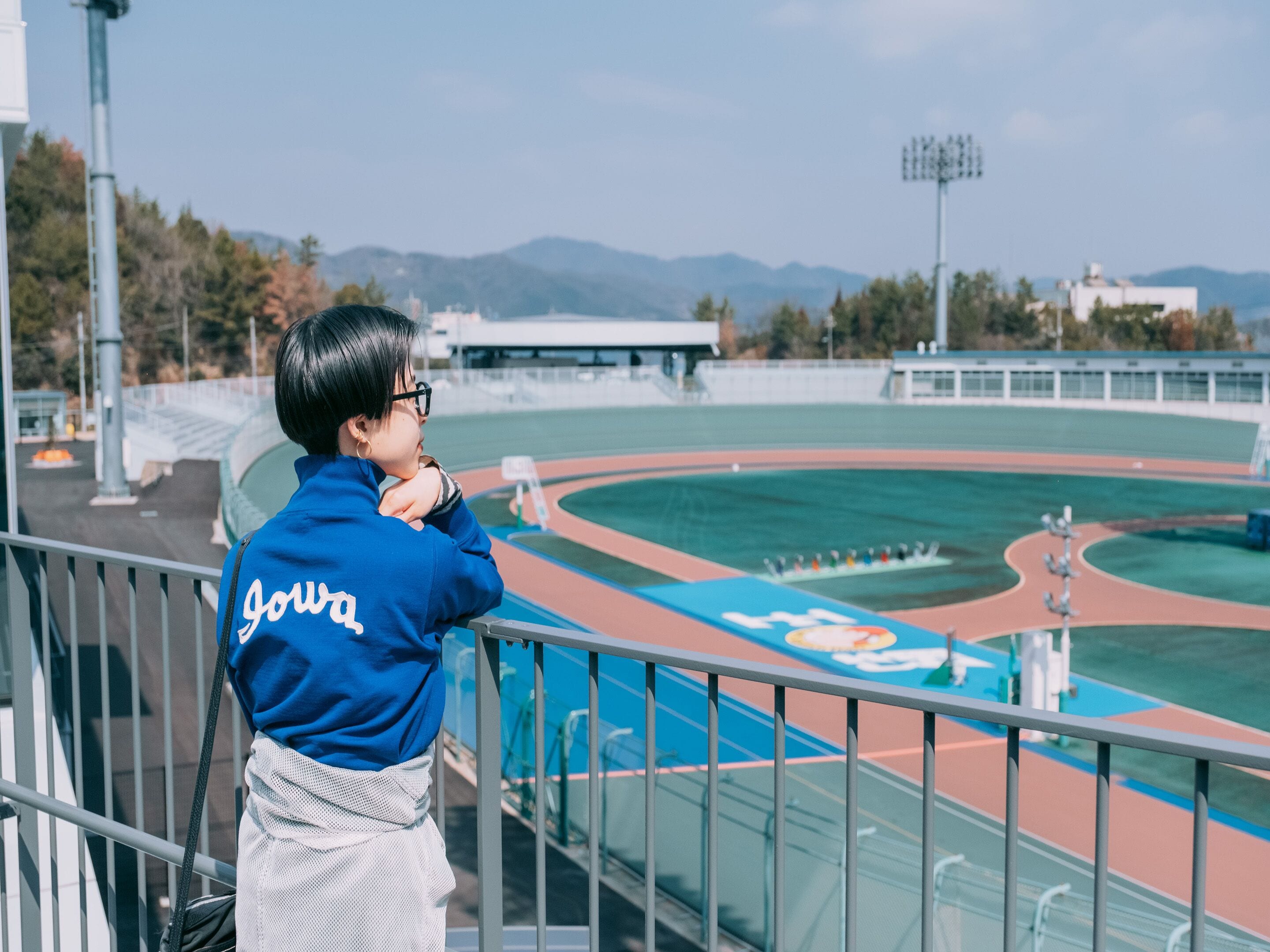 A person in a blue jacket with "Joya" on the back, overlooking a sports track with a blue and red running path, surrounded by trees and distant mountains.