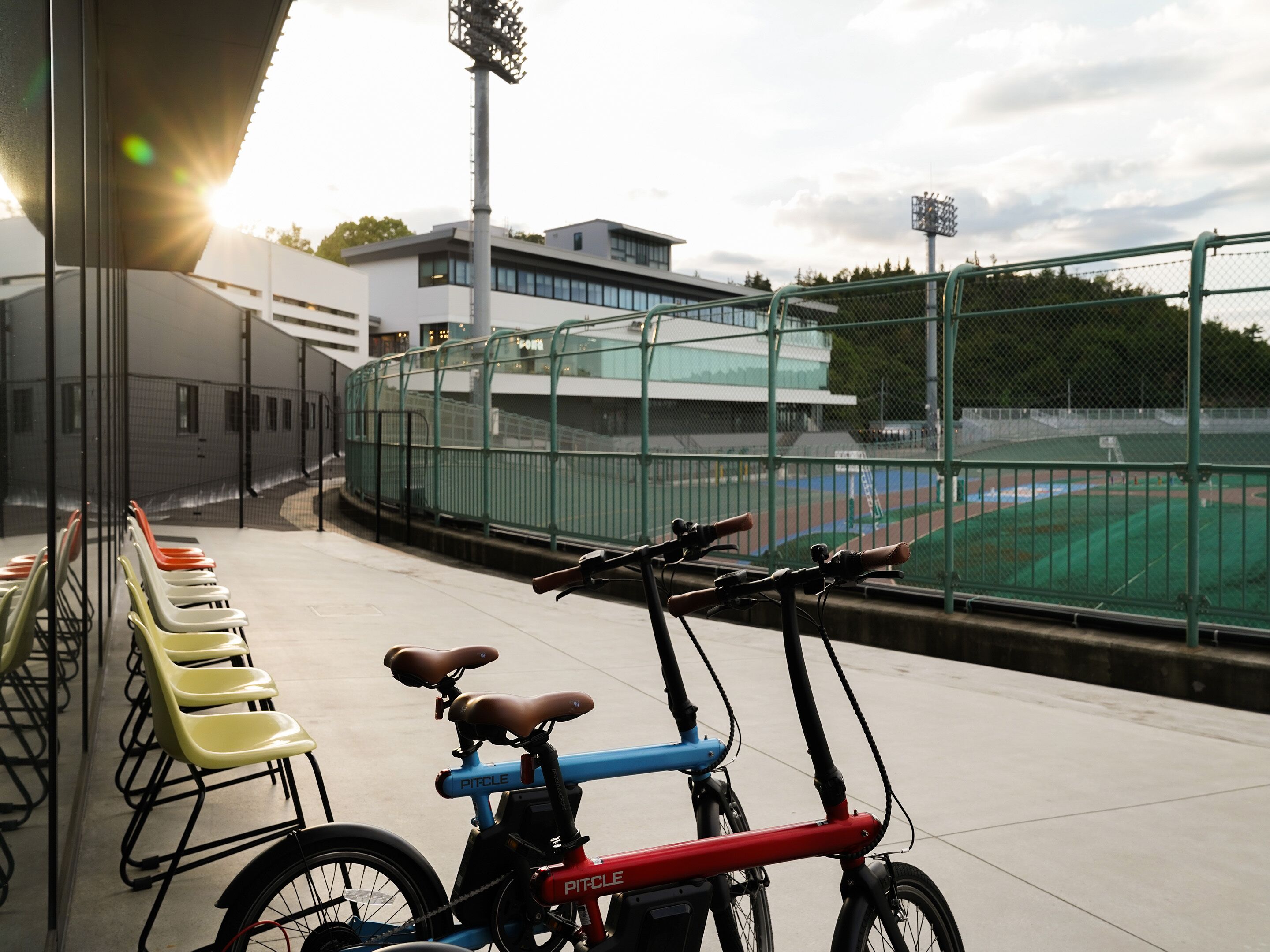 A row of yellow chairs and a red and blue bicycle parked near a sports field.