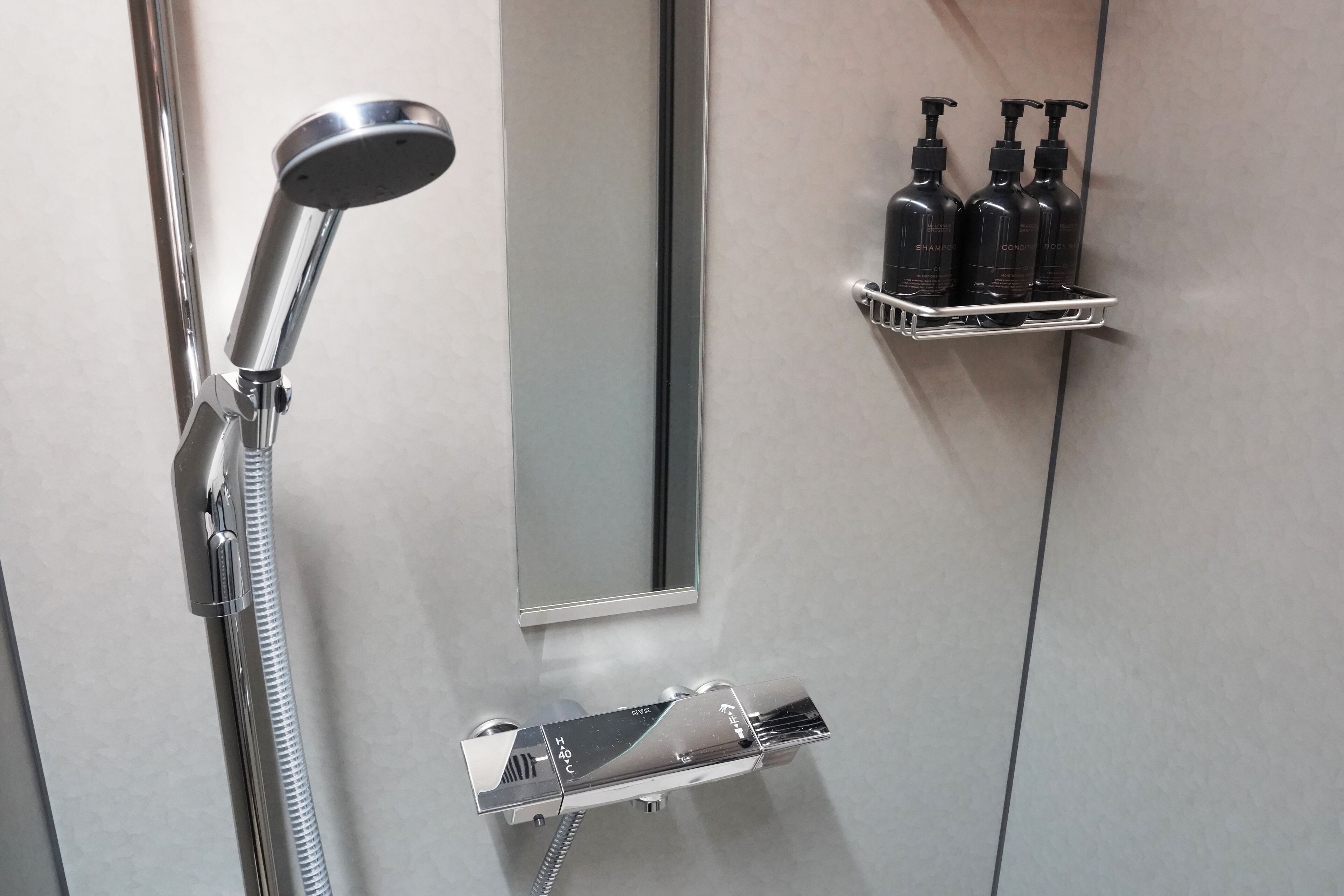 A bathroom with a shower, a mirror, and a shelf with three bottles of shampoo and conditioner.