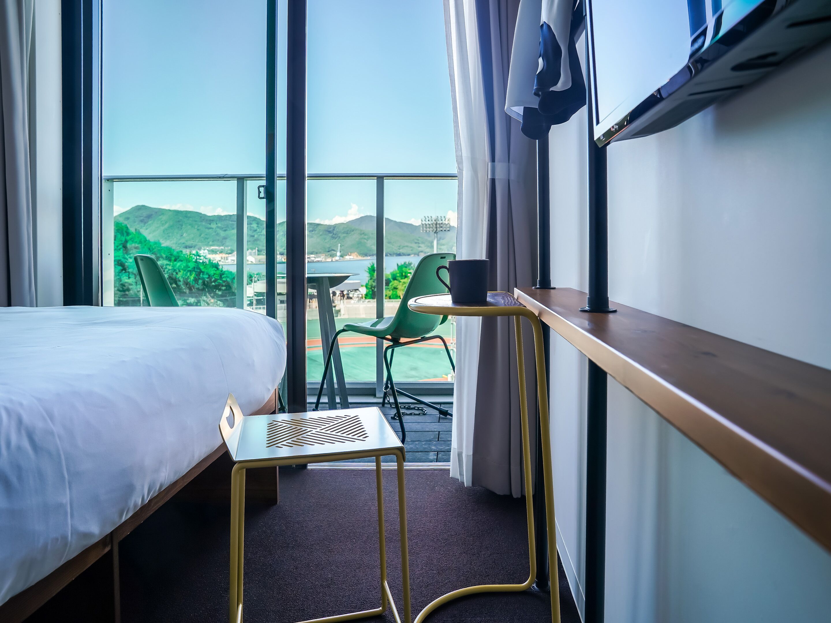 A hotel room with a bed, a small table, a chair, and a view of a balcony with greenery and a distant landscape.