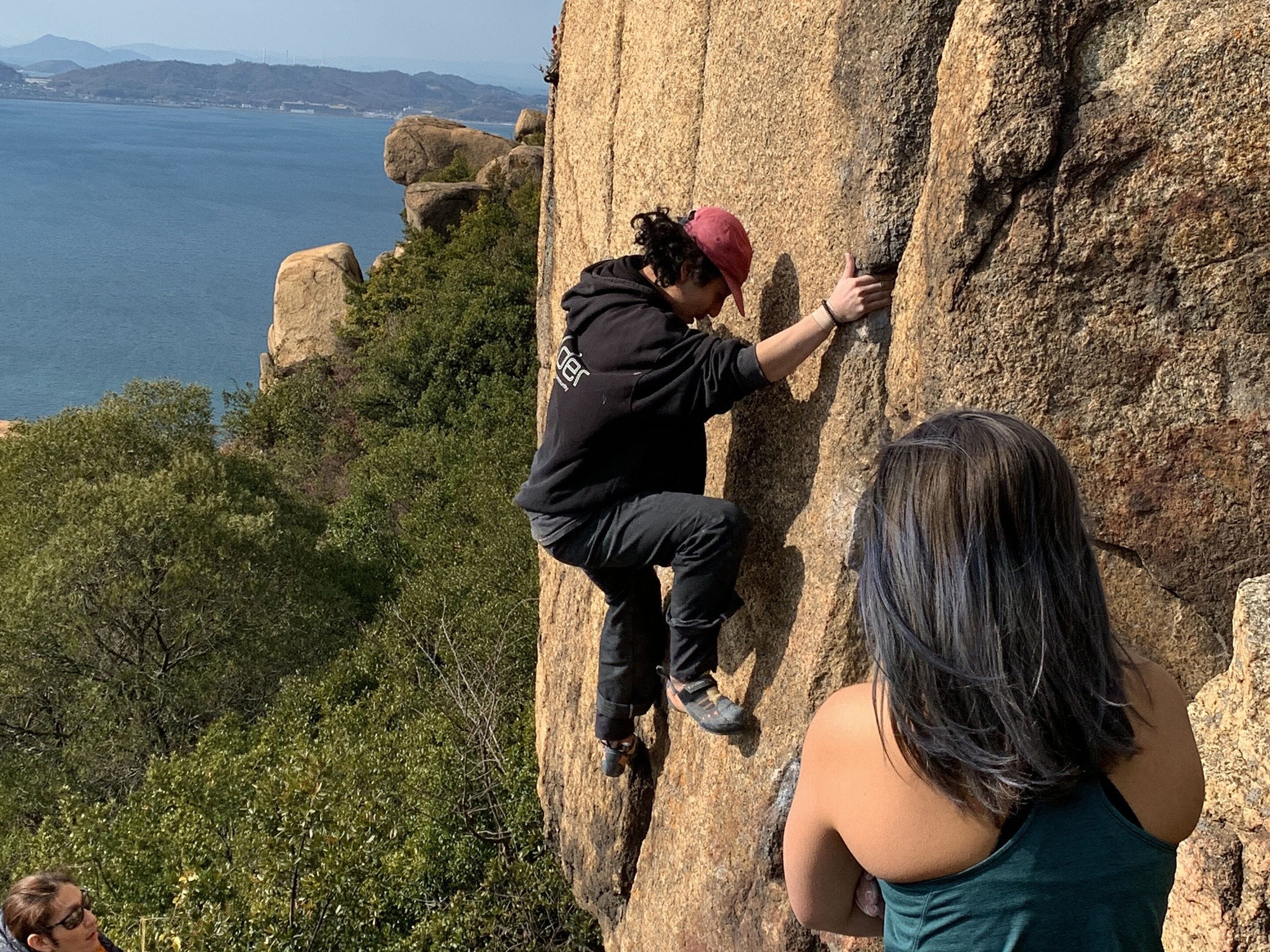 A person climbing a rock face with a coastal view in the background.
