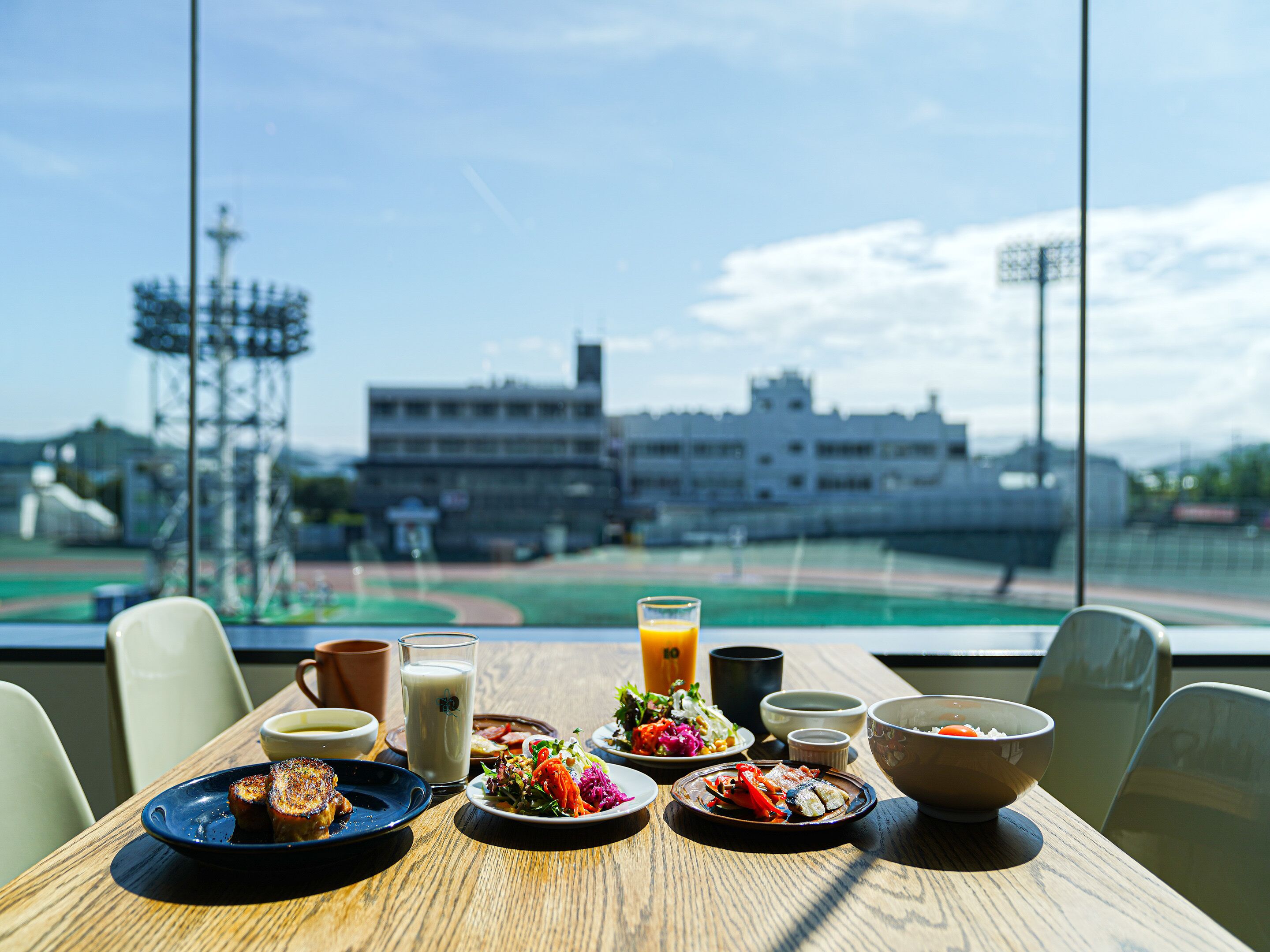 A table with various dishes, a glass of orange juice, and a view of a sports field and buildings in the background.