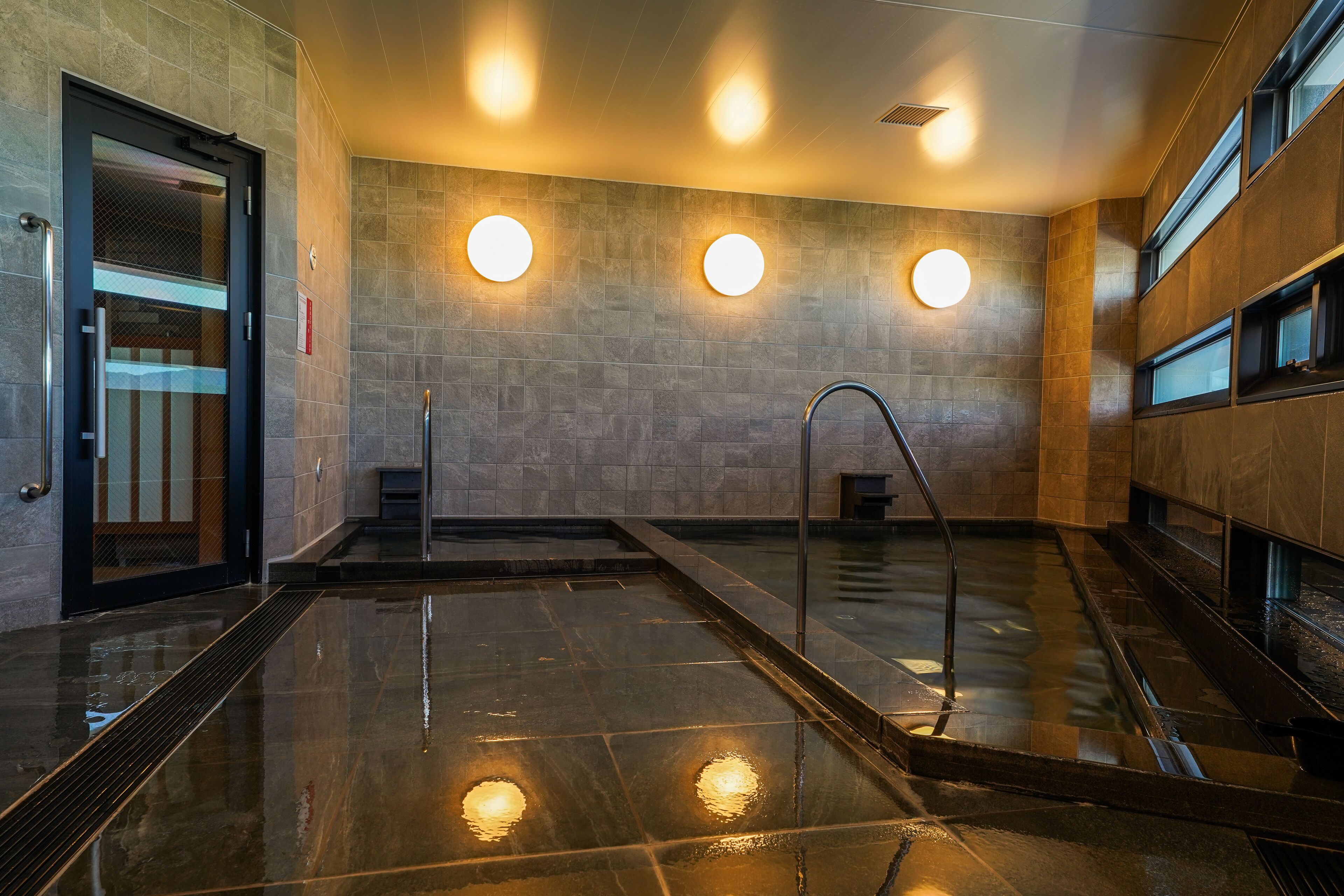 Un área de piscina interior moderna con entrada por puerta de vidrio, un letrero de piso mojado y tres focos redondos en la pared.