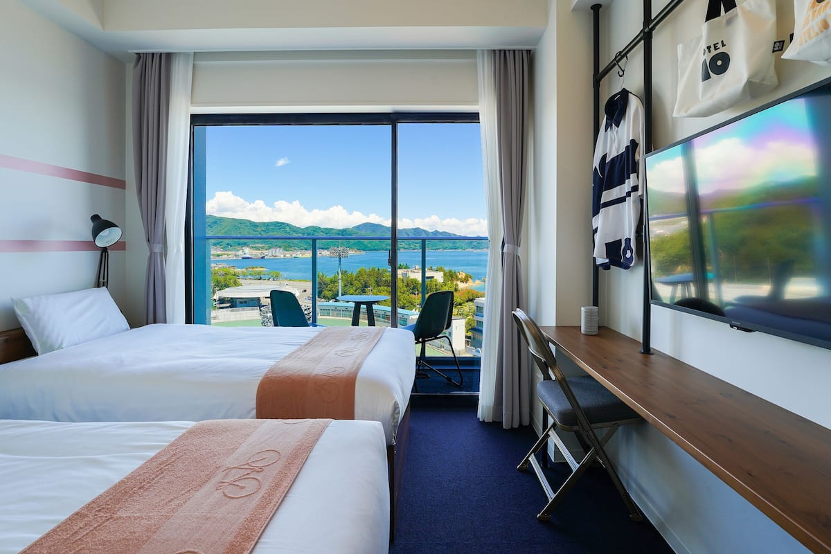 A hotel room with two beds, a desk, a TV, and a view of the sea and mountains.