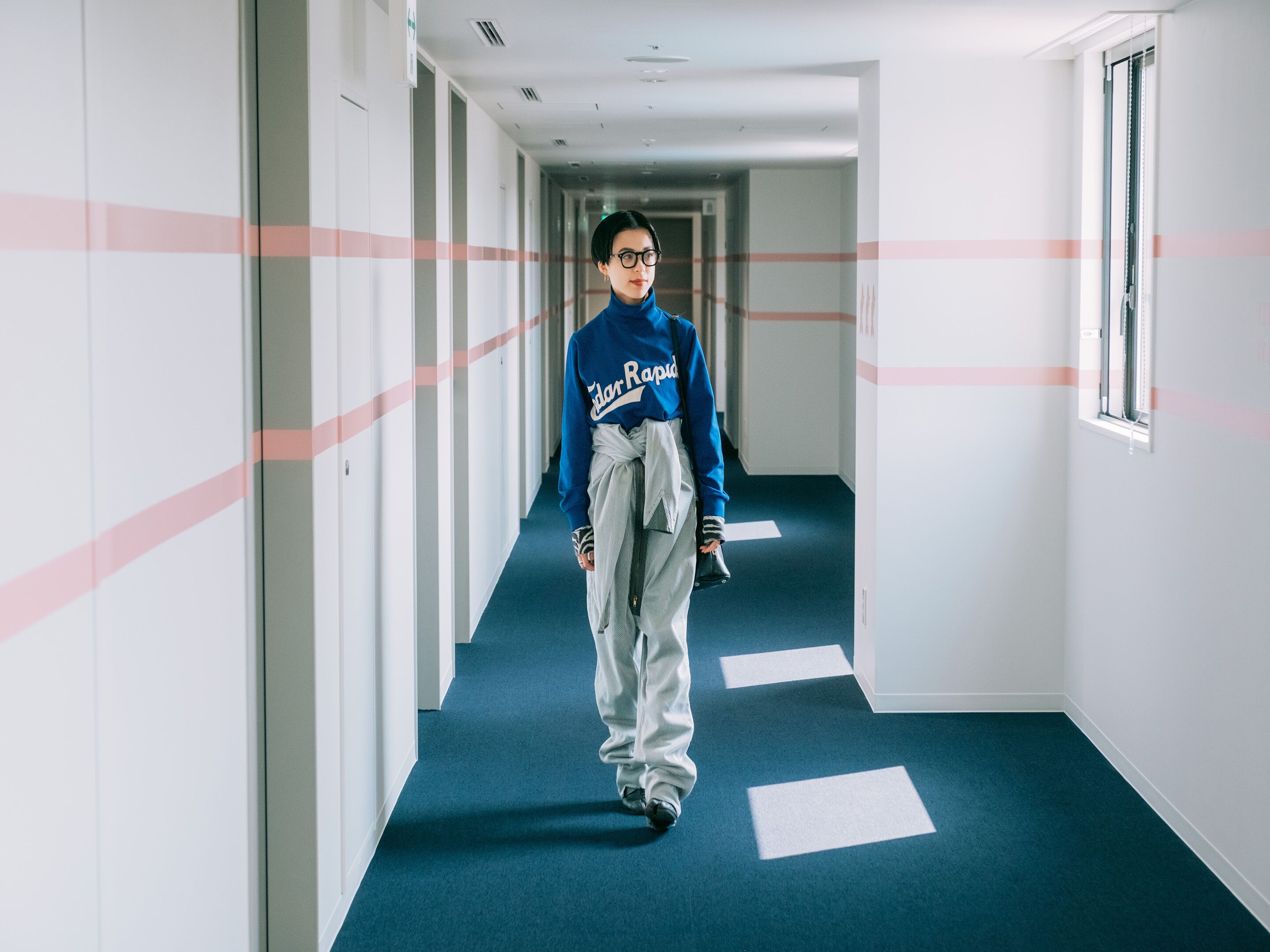 Una persona caminando por un pasillo con alfombra azul y paredes blancas, con una ventana a la derecha.