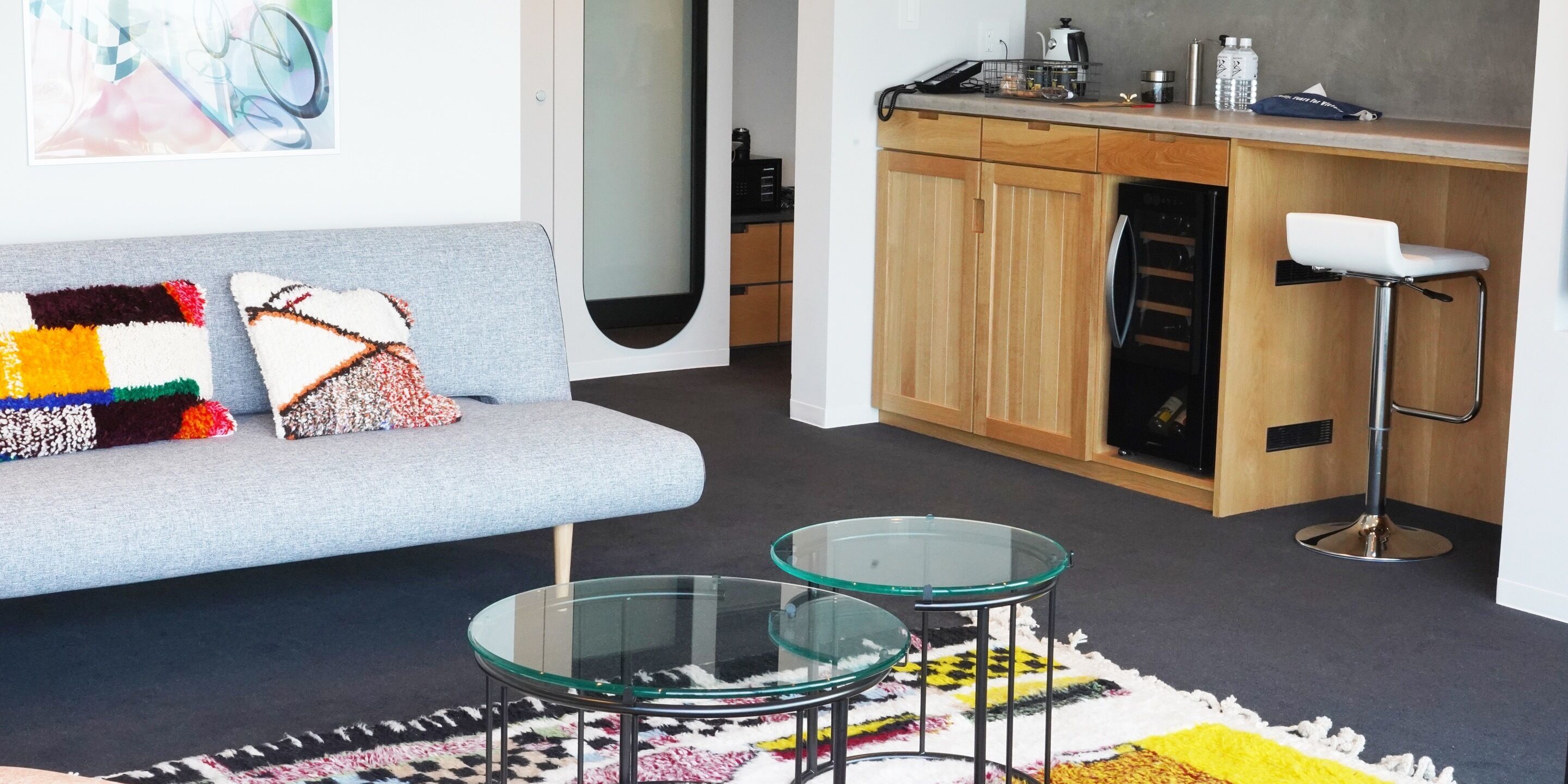 A modern living room with a grey sofa, two glass tables, and a colorful rug.