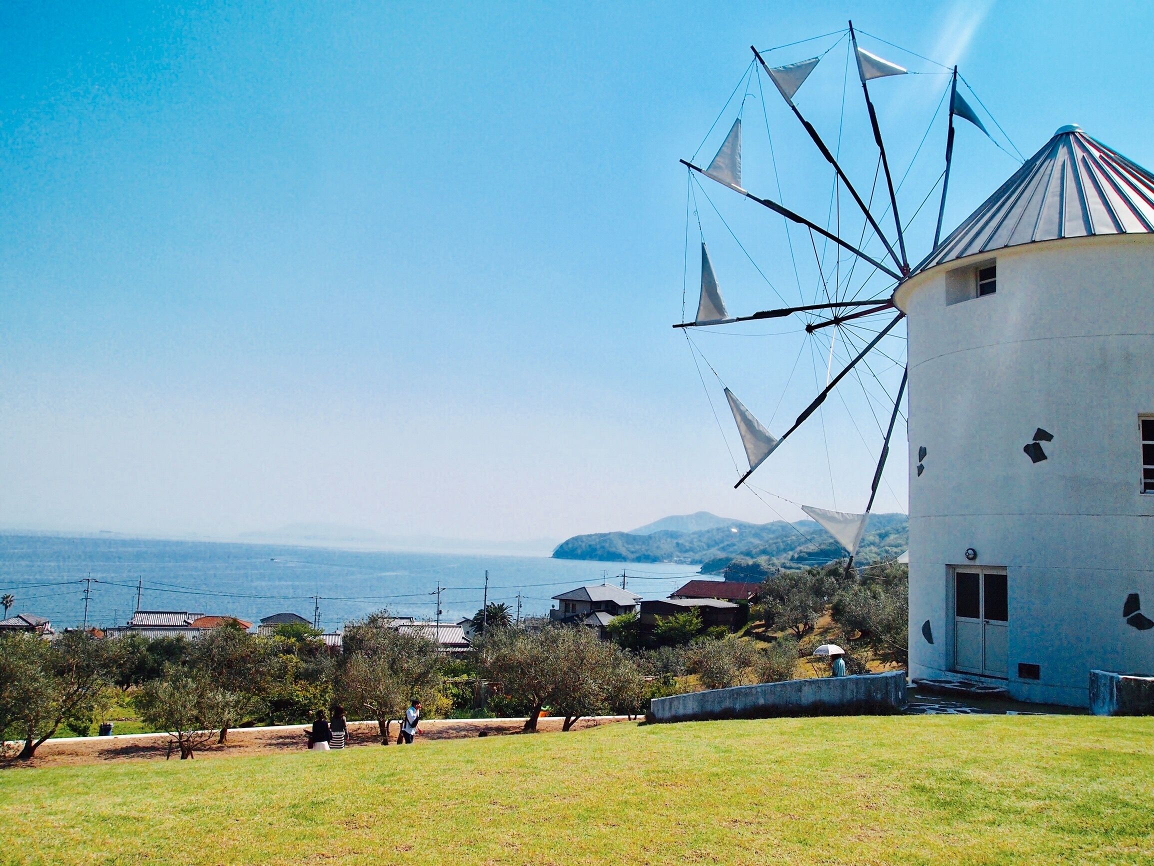 Un moulin à vent, avec un bâtiment blanc à proximité, une zone herbeuse et des maisons en arrière-plan.