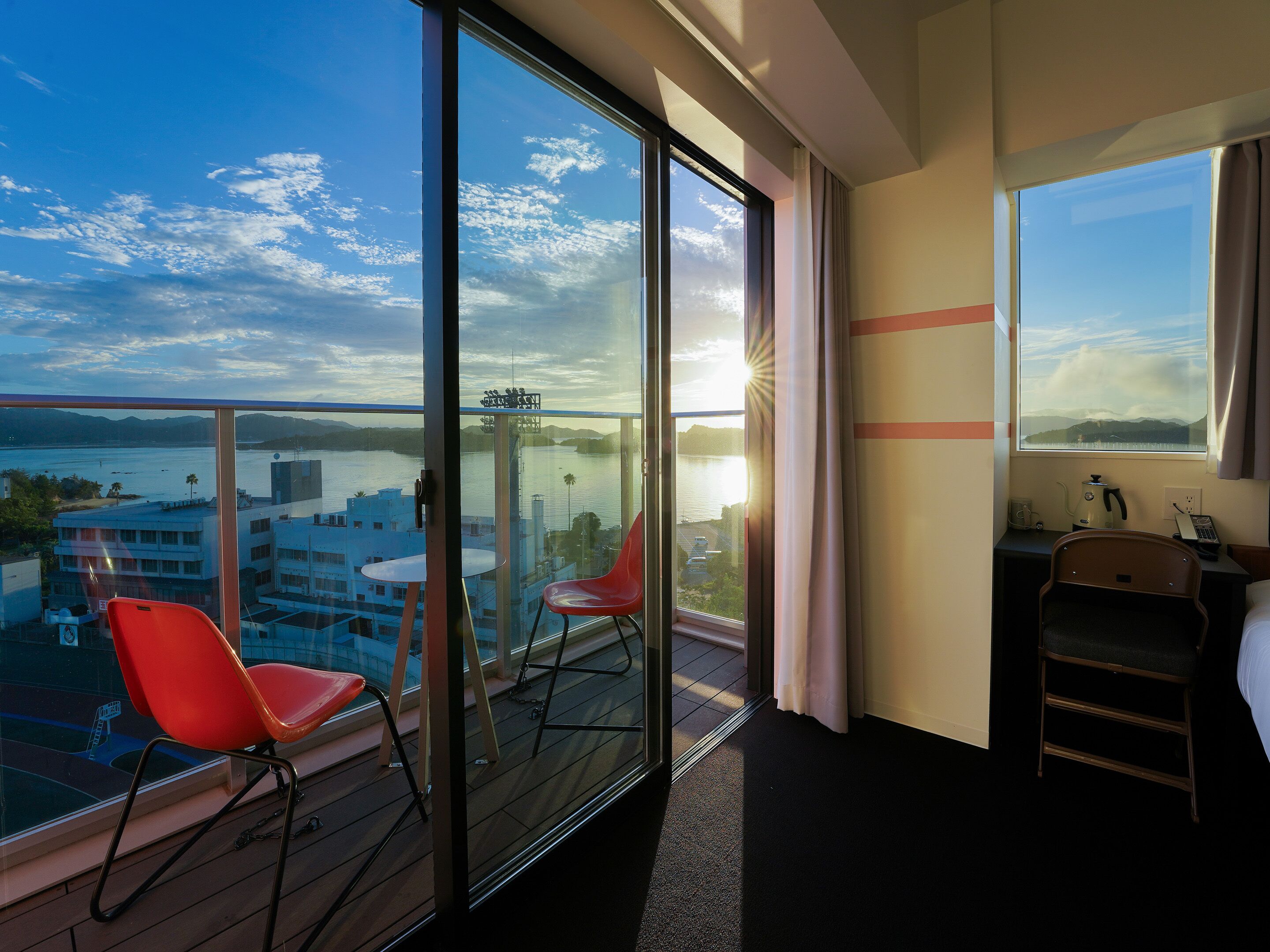 Habitación de hotel con balcón grande, una silla roja, una mesita y vistas a un cuerpo de agua y edificios.