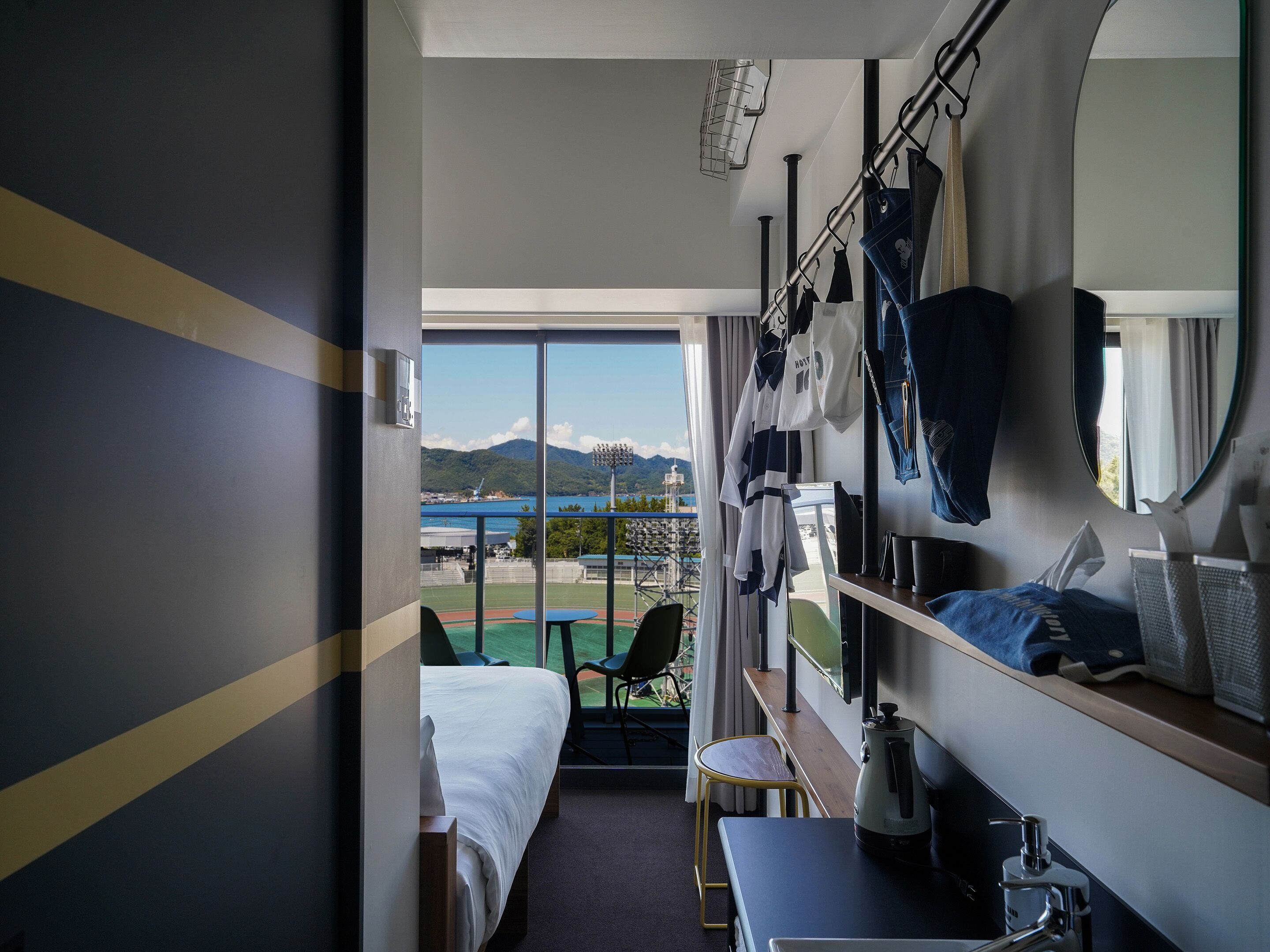 A hotel room with a bed, a desk, a chair, and a view of a sports field and mountains.