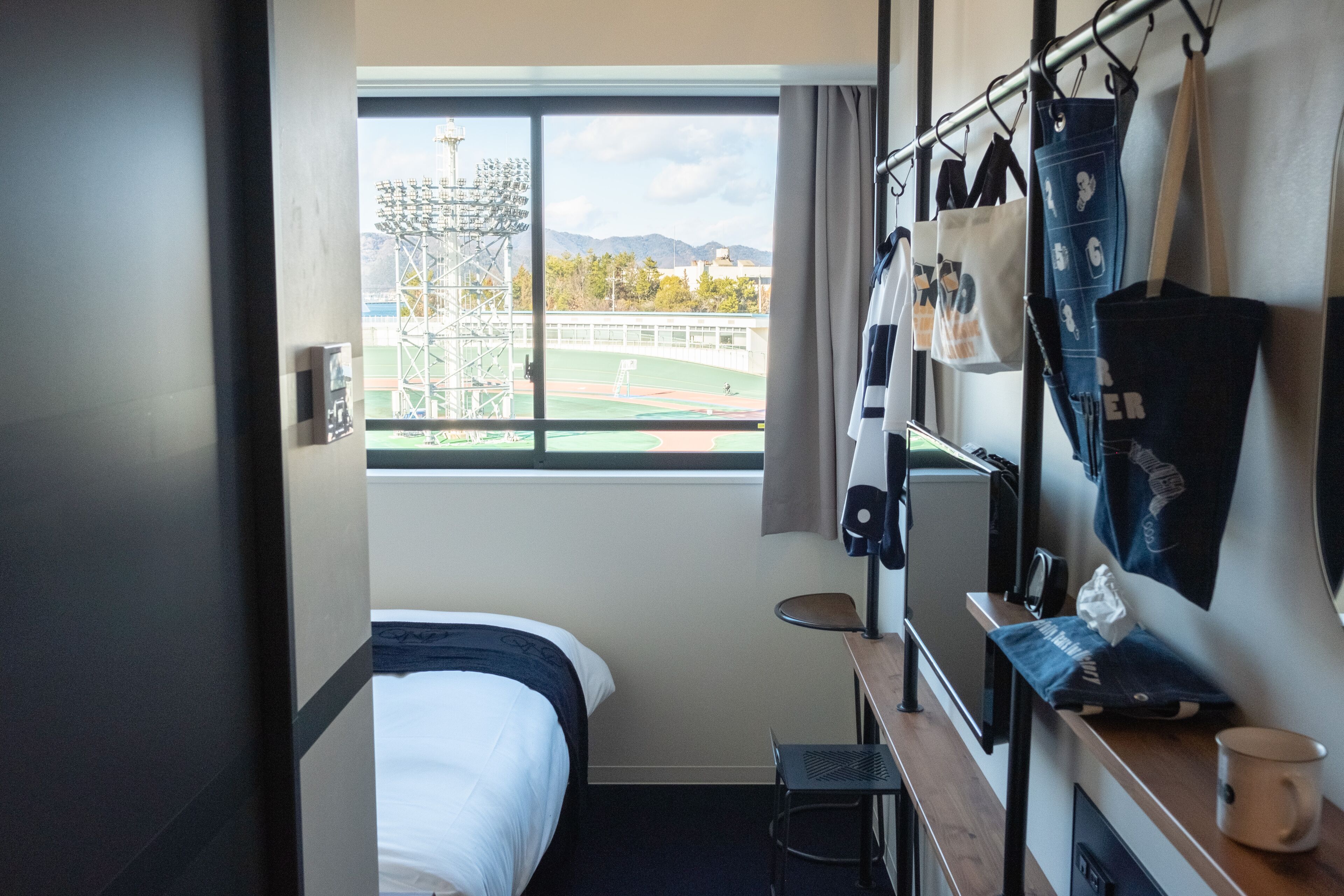 A hotel room with a bed, a desk, a chair, a window with a view of a sports field, and a hanging bag rack.