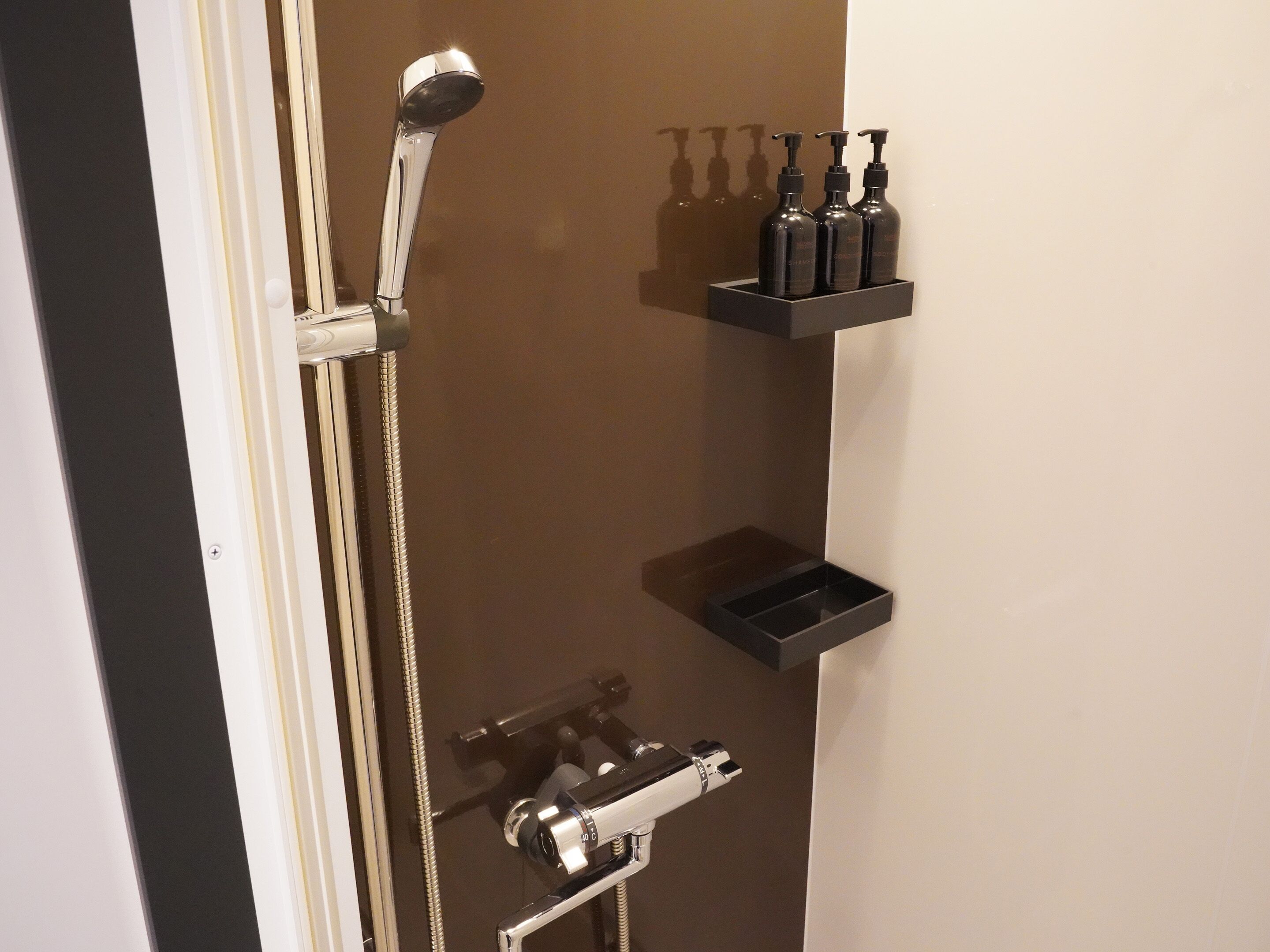 A shower with a handheld showerhead, a wall-mounted faucet, and a handheld shower attachment. There are three black bottles on a black shelf.