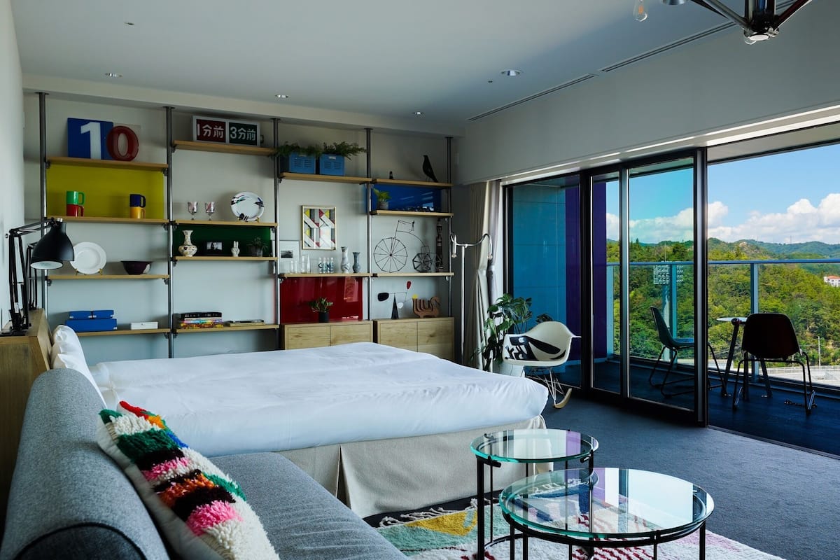 A modern bedroom with a large bed, a glass table, and a balcony with a city view.