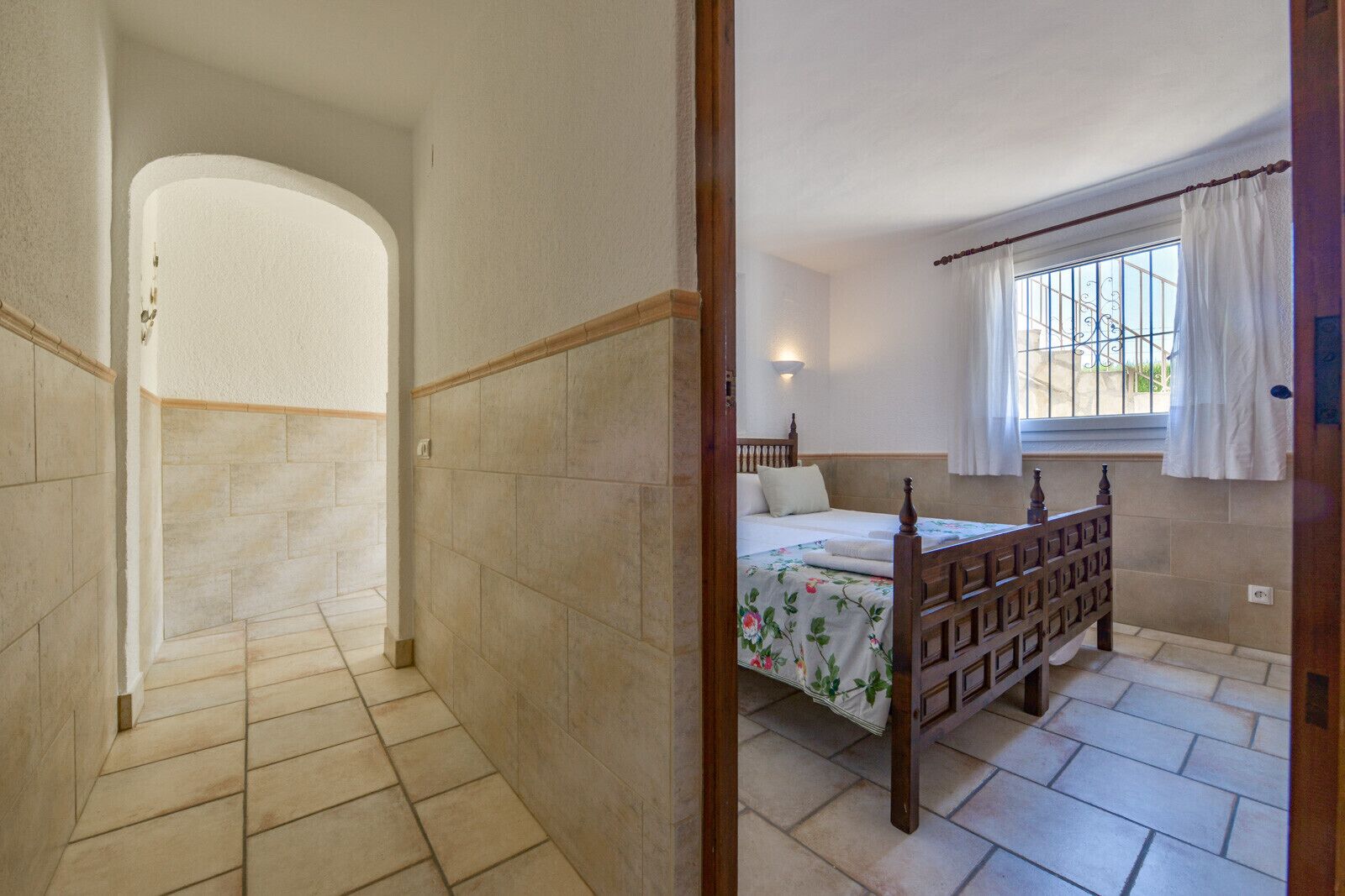 A bedroom with a wooden bed, a window with curtains, and a tiled floor.