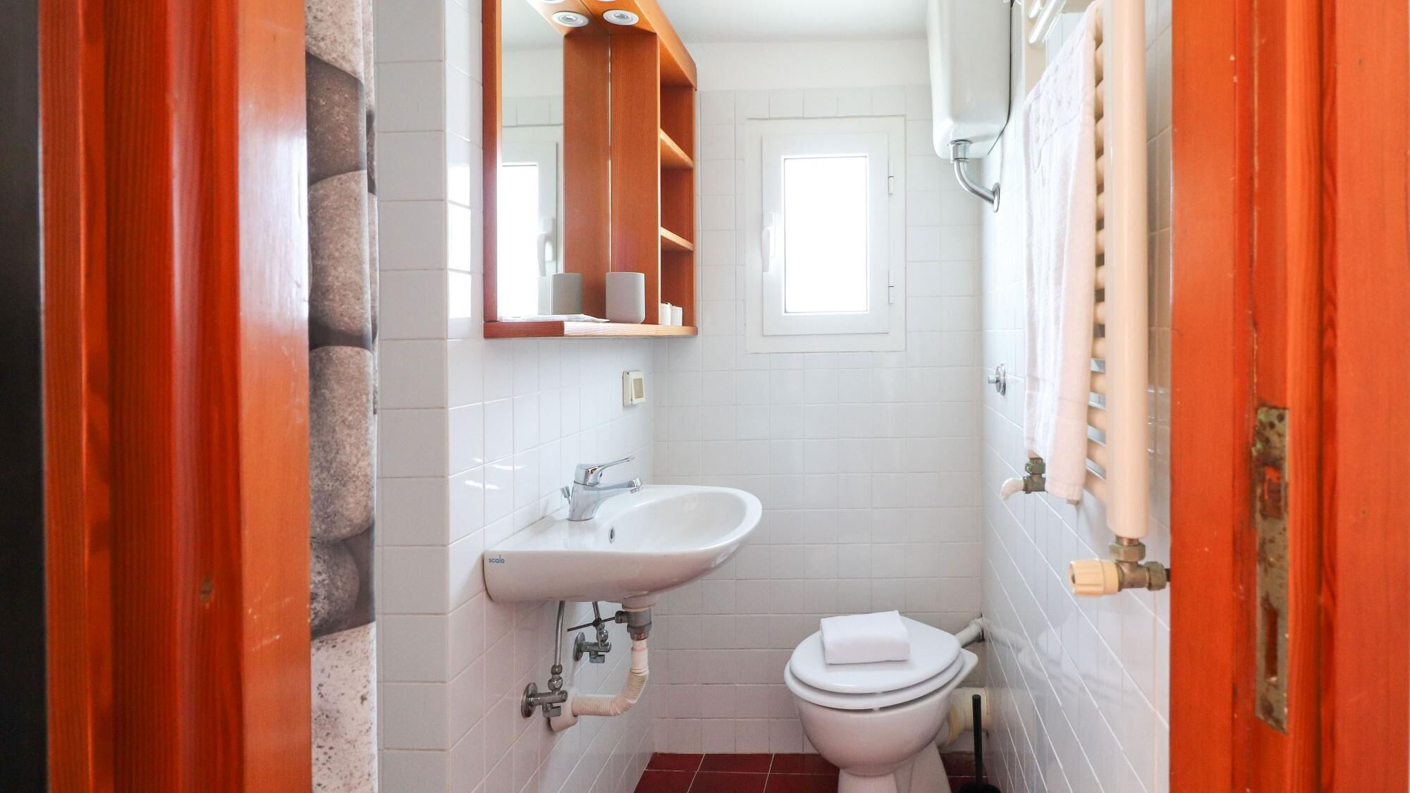 A small bathroom with a white sink, a toilet, and a mirror above the sink. There is a window above the toilet and a towel rack on the wall.