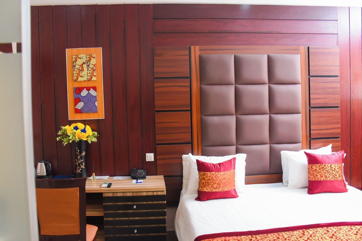 A hotel room with a wooden headboard, a desk with a telephone, a vase of flowers, and a bed with patterned pillows.
