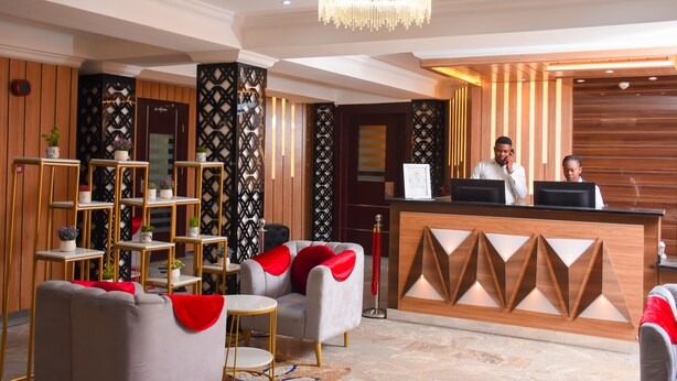 A hotel reception area with a wooden reception desk, two receptionists, a chandelier, and a seating area with sofas and a small round table. - Golden Tulip Garden City Hotel - Rivotel (Port Harcourt)