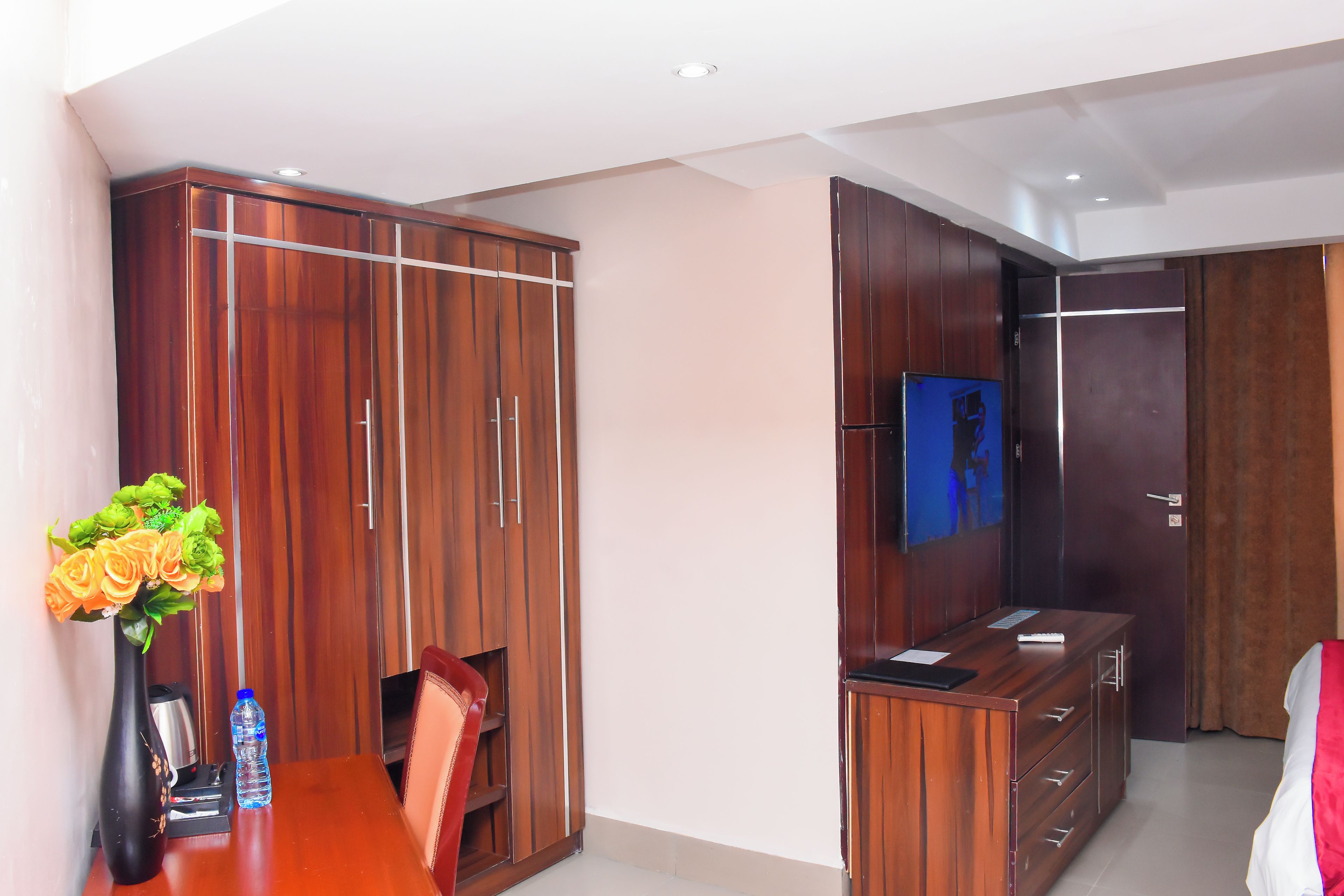 A hotel room with a wooden wardrobe, a television mounted on the wall, a desk with a vase of flowers, and a water bottle.