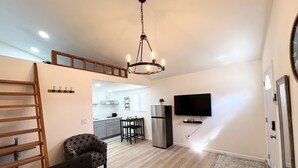 A modern living room with a sofa, a patterned ottoman, a wooden ladder, a dining area with a table and chairs, a kitchen with a refrigerator and a TV mounted on the wall.
