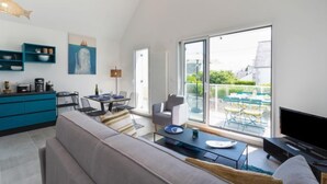 A modern living room with a sofa, a dining table, and a TV. There is a kitchen area with blue cabinets and a coffee machine. A large window offers a view of the outdoors. - Le Clos Moguer by Interhome (Quiberon)
