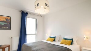 A bedroom with a bed, a bedside table, a lamp, a window with curtains, and a wall-mounted light fixture. - Le Clos Moguer by Interhome (Quiberon)