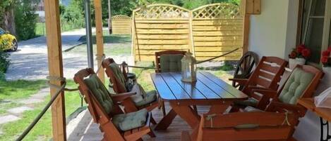 A covered patio with a wooden table and chairs, a lantern, and a view of a wooden fence and greenery.