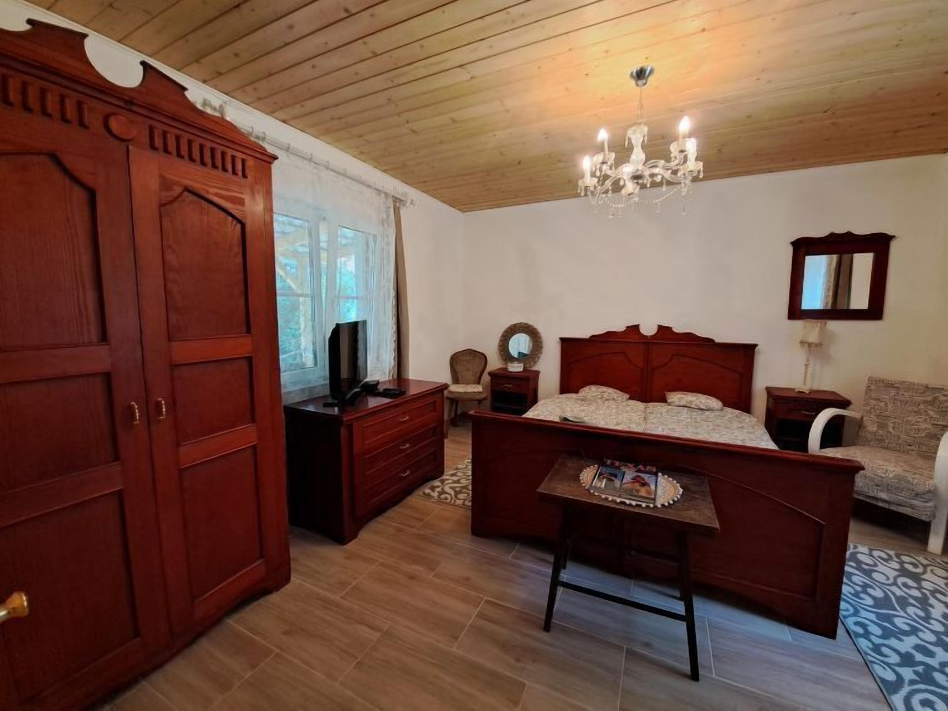 Un dormitorio con techo de madera, un candelabro, una cama con colcha estampada, un aparador de madera, un televisor y una ventana con cortinas.