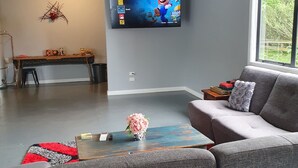 A modern living room with a grey sofa, a coffee table with a vase of flowers, a flat-screen TV mounted on the wall, and a large window offering a view of greenery outside. - The Red Shed
Large modern studio on two peaceful acres, two minutes from CBD (Matamata)