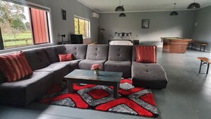 A modern living room with a grey sofa, a coffee table, and a pool table. There are pendant lights and a large window with red shutters. - The Red Shed
Large modern studio on two peaceful acres, two minutes from CBD (Matamata)