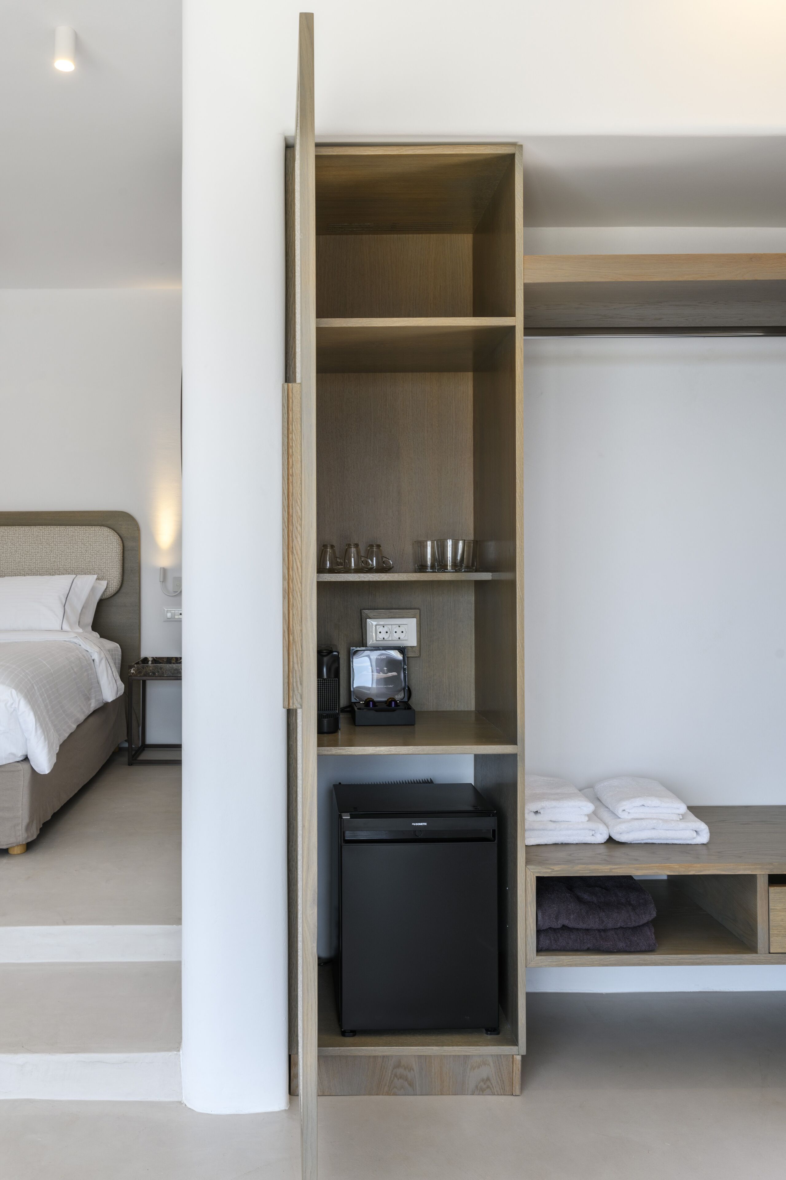 A hotel room with a bed, a wardrobe with shelves, a black refrigerator, and a nightstand with a lamp.
