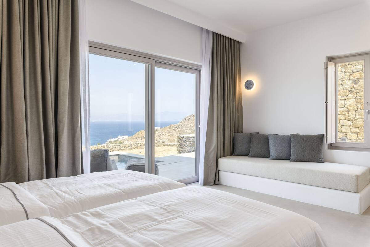 A hotel room with two beds, a sofa, a stone wall, and a view of the sea.