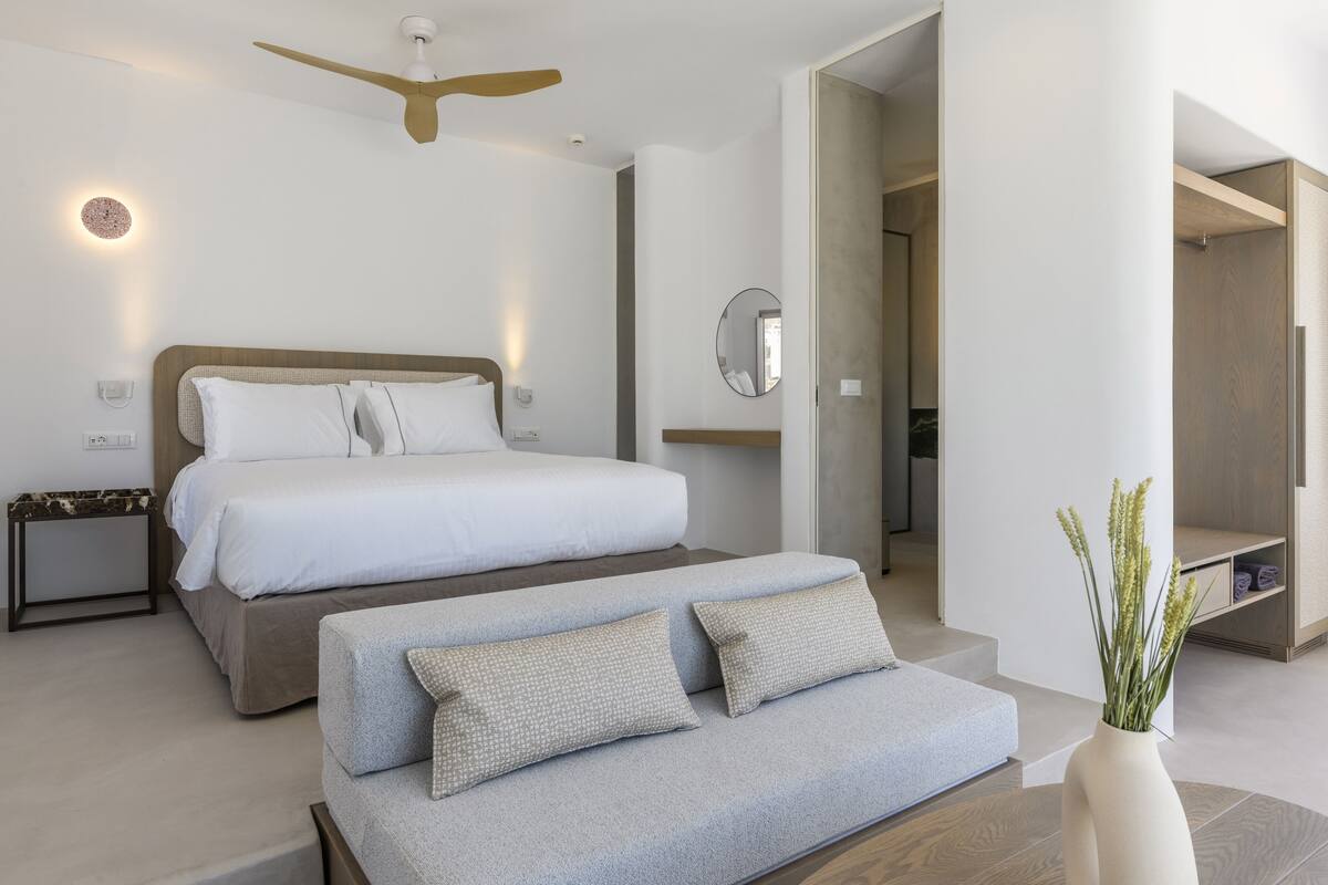 A modern bedroom with a large bed, a sofa, a ceiling fan, and a mirror.