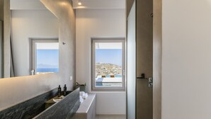 Un baño moderno con encimera de mármol, dos ventanas con vistas al paisaje costero y una puerta que da a otra habitación.