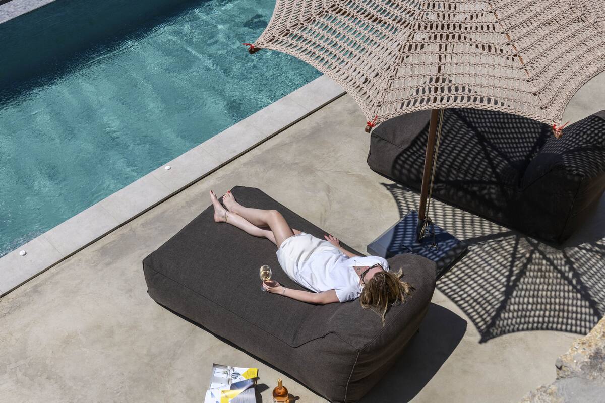 A person relaxing on a dark-colored lounge chair by a poolside, with a woven umbrella providing shade.