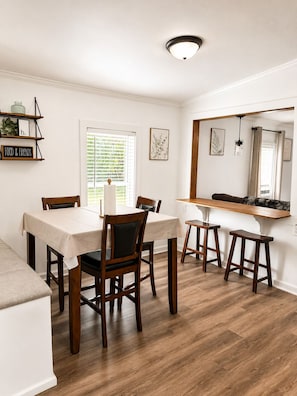 A dining area with a table, chairs, and a bench. There is a window with blinds, a shelf with decorative items, and a mirror above the counter.