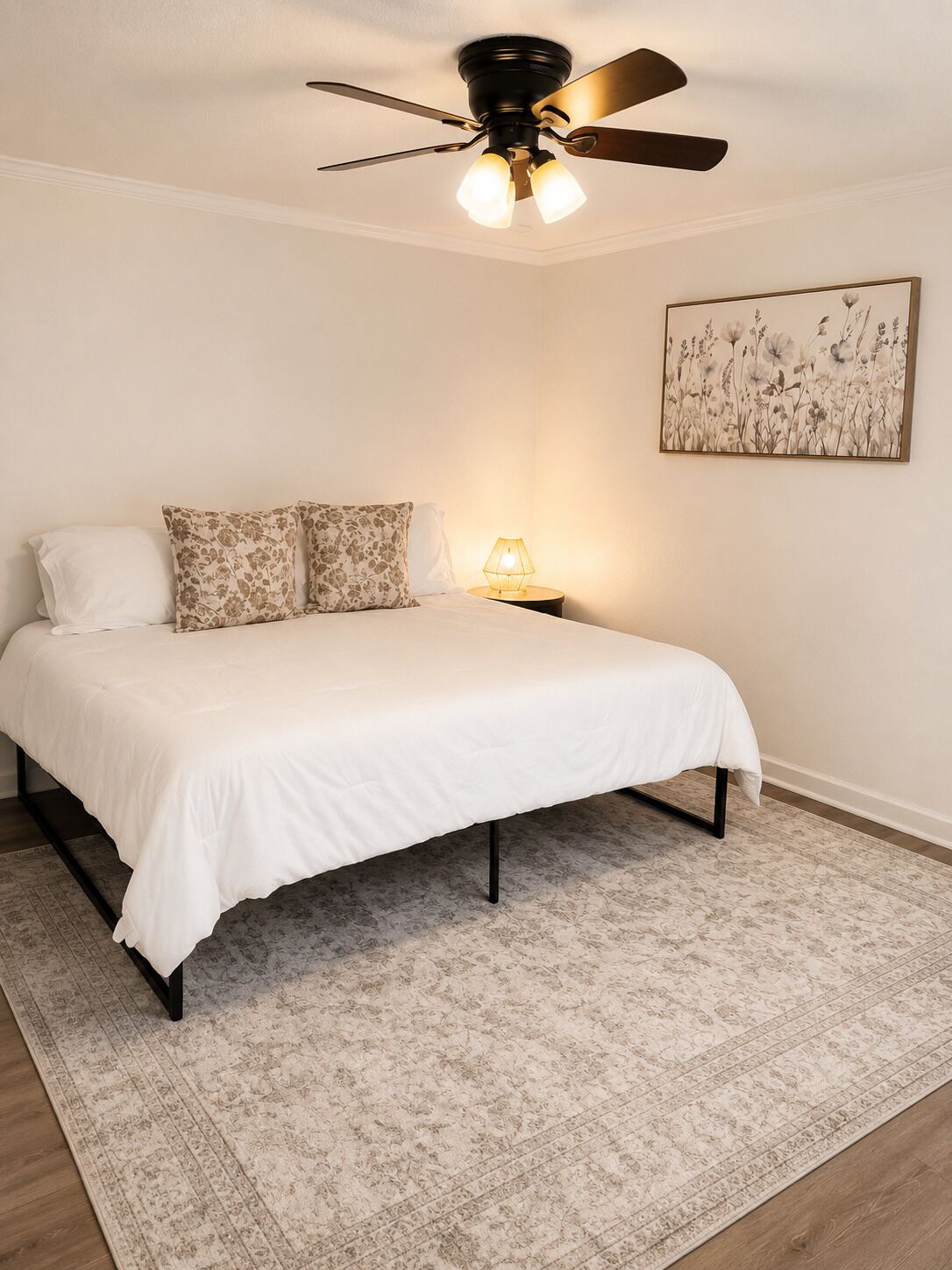 Un dormitorio con cama, mesita de noche, lámpara, ventilador de techo y una obra de arte floral en la pared.