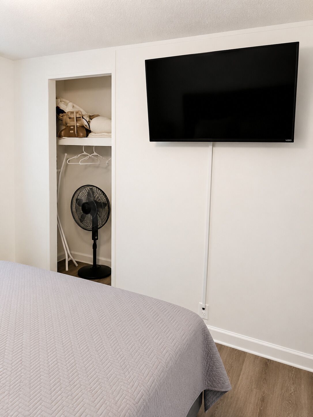 Une chambre à coucher avec un lit, un téléviseur fixé au mur, un ventilateur et une armoire où sont suspendus des vêtements.