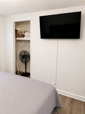 A bedroom with a bed, a TV mounted on the wall, a fan, and a closet with hanging clothes.
