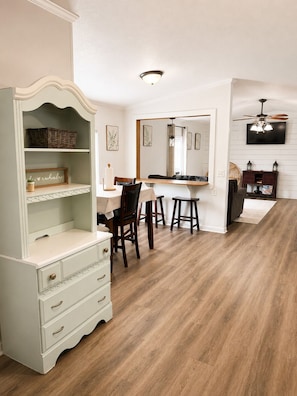A dining area with a white table, chairs, and a wooden floor. There is a white dresser with drawers and a mirror above it.