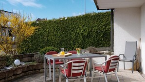 Eine Terrasse im Freien mit Tisch und Stühlen, ein Hecken im Hintergrund und eine Steinmauer.