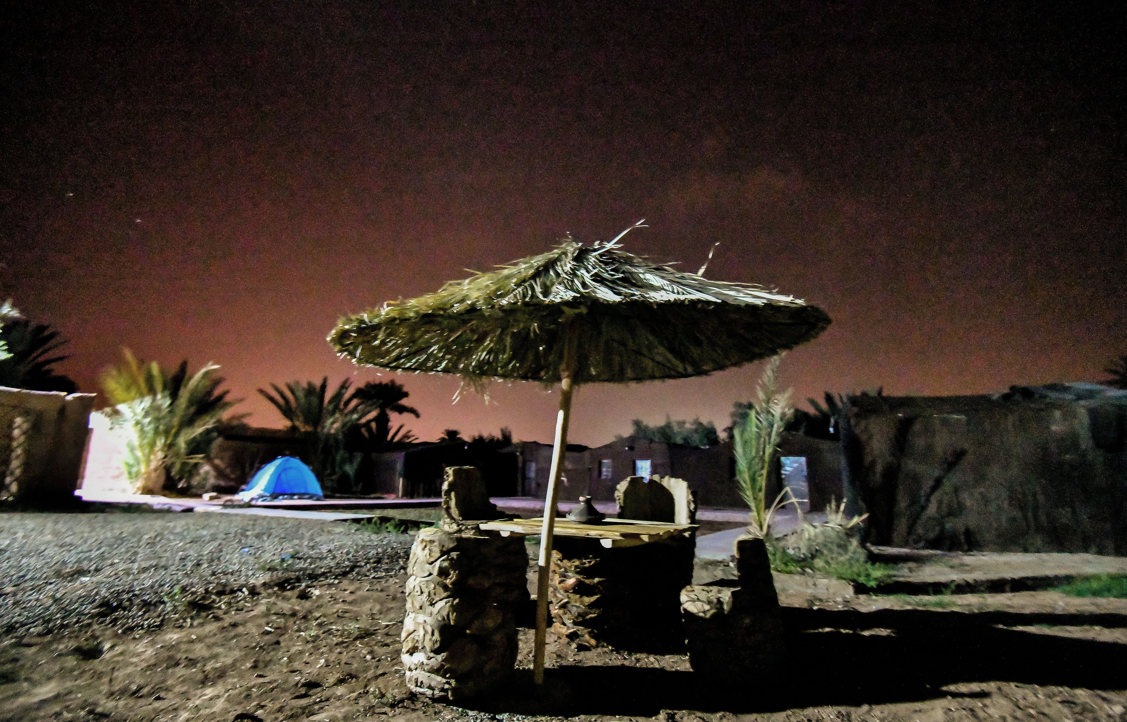 Een rustieke buitenomgeving met een rieten parasol, stenen krukken en een blauwe tent.