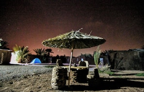A rustic outdoor setting with a thatched umbrella table, stone stools, and a blue tent.