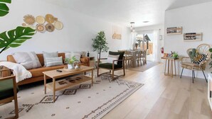 Ein modernes Wohnzimmer mit einer Couch, einem Couchtisch und Sesseln. Es gibt einen Essbereich mit Tisch und Stühlen. Das Zimmer verfügt über ein großes Fenster und eine Pflanze.