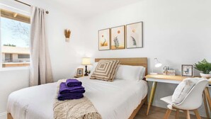 Ein modernes Schlafzimmer mit einem großen Bett, einem Schreibtisch und einem Stuhl. An den Wänden hängen Bilder in Rahmen, auf dem Schreibtisch steht eine Lampe und es gibt ein Fenster mit Vorhängen.