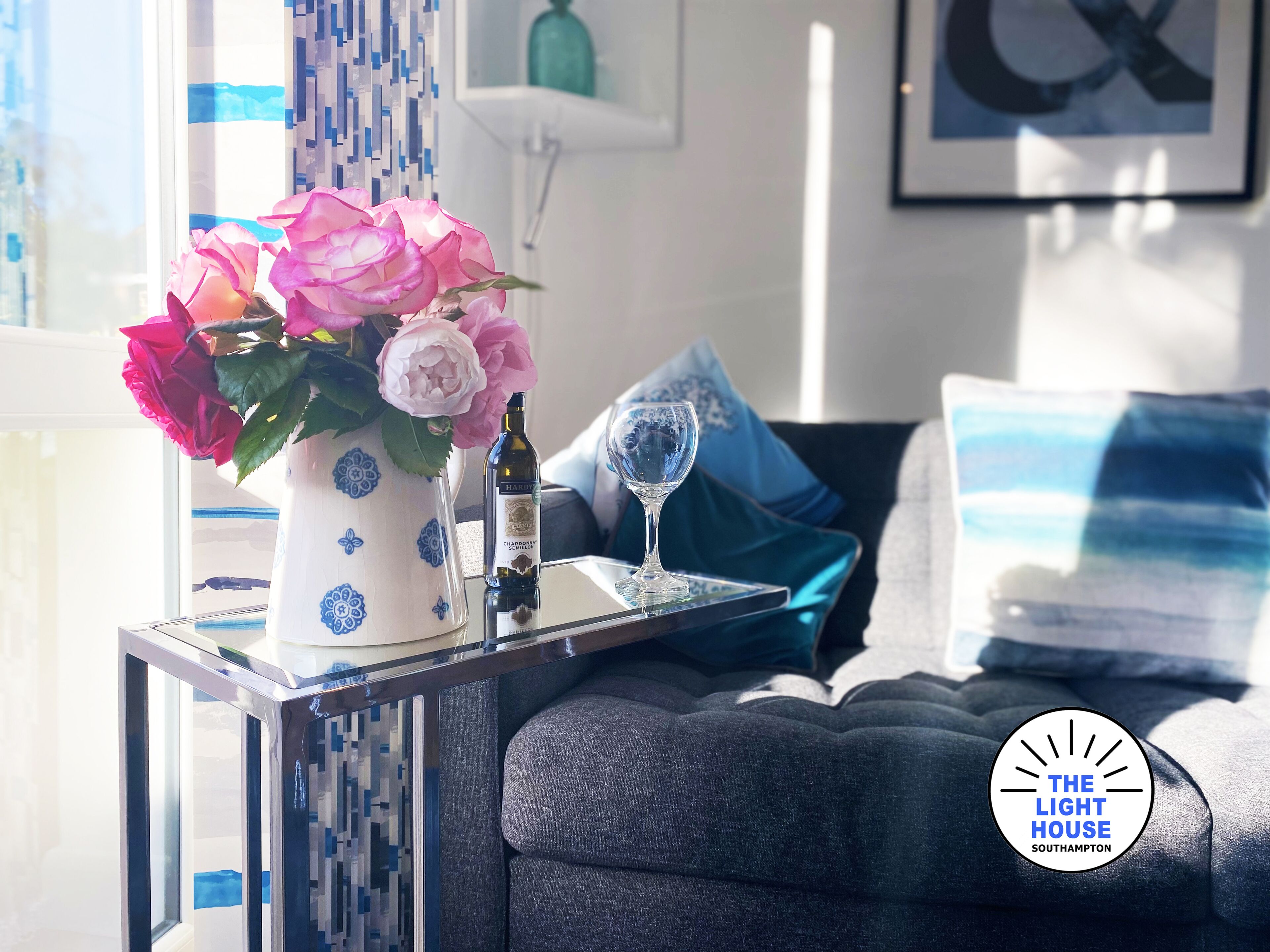 A modern living room with a sofa, decorative pillows, a vase with flowers, and a wine bottle on a side table.