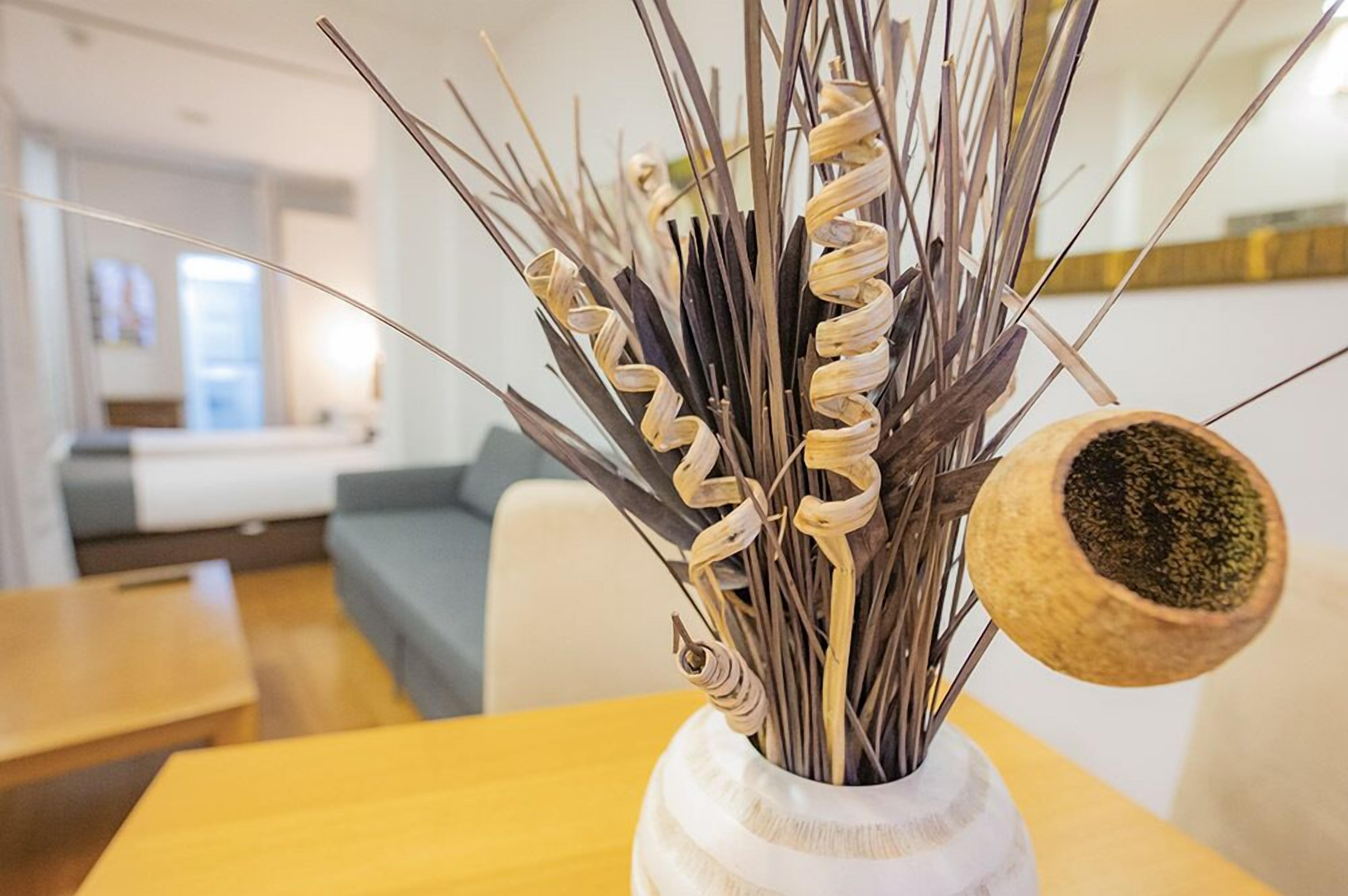 A room with a wooden table, a white vase with dried plants, and a grey sofa.